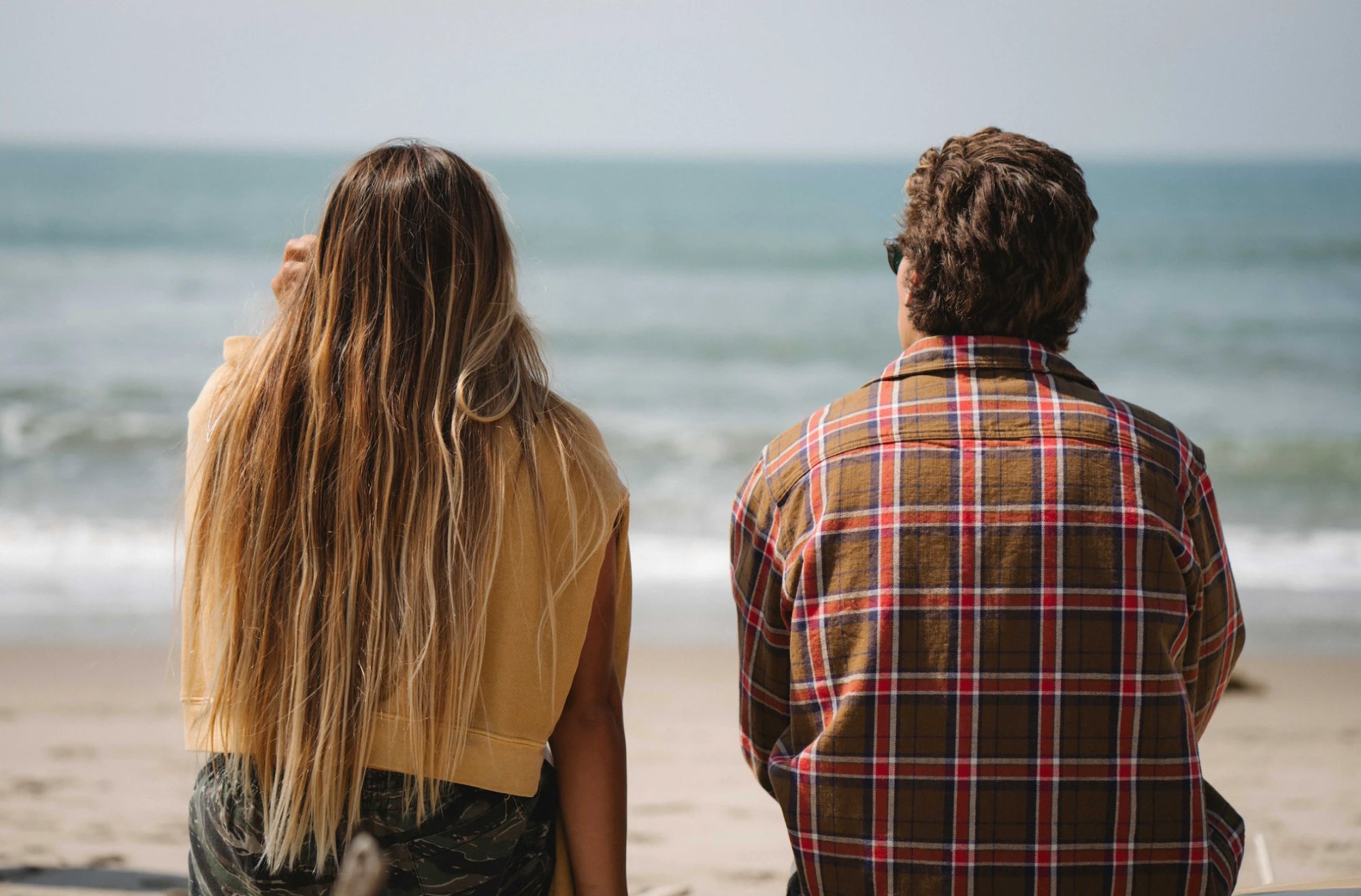 Woman and man sitting in front of water at beach wearing Ariko LA Clothing